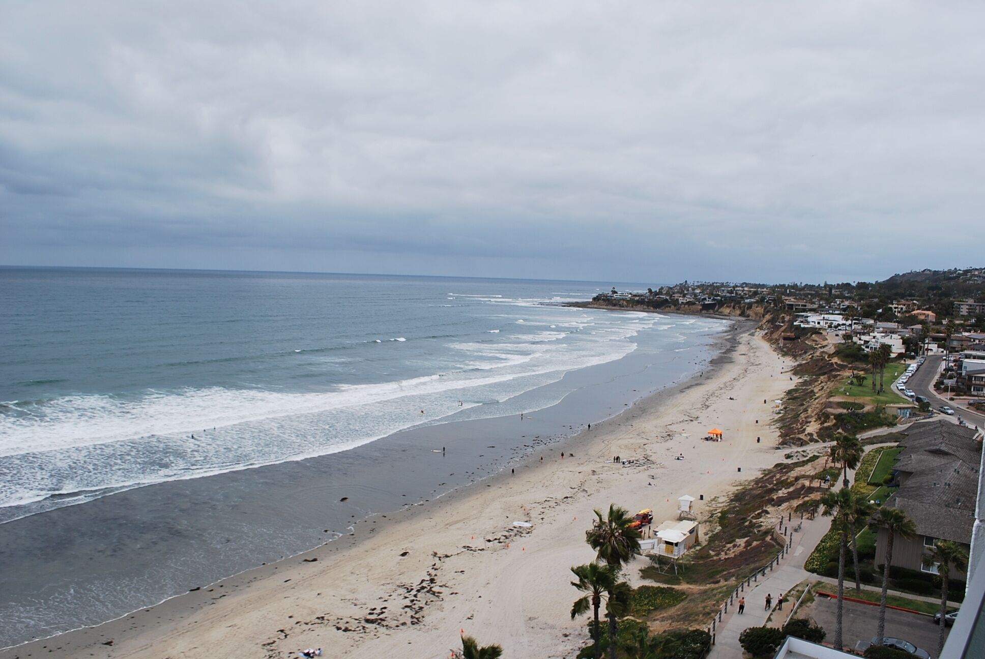 A coastal beach with a wide sandy shore, a few palm trees, and a residential area in the background.