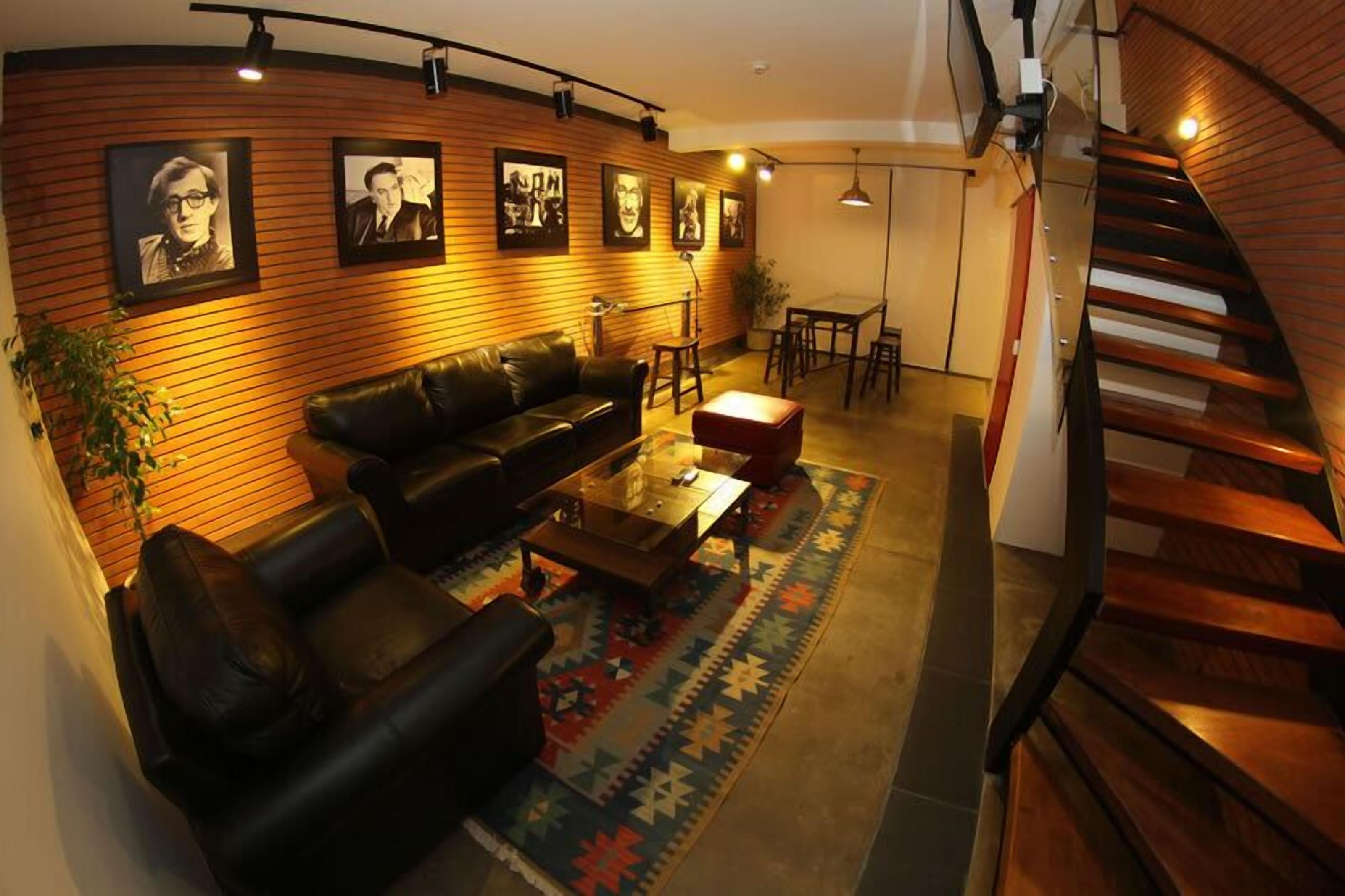 A modern living room with a black leather sofa, a glass coffee table, and a colorful rug. There are framed pictures on the wall, a staircase, and a dining area with a table and chairs.