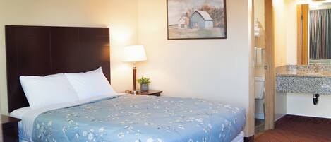 A hotel room with a bed, a nightstand, a lamp, a painting, a bathroom with a sink, and a mirror.