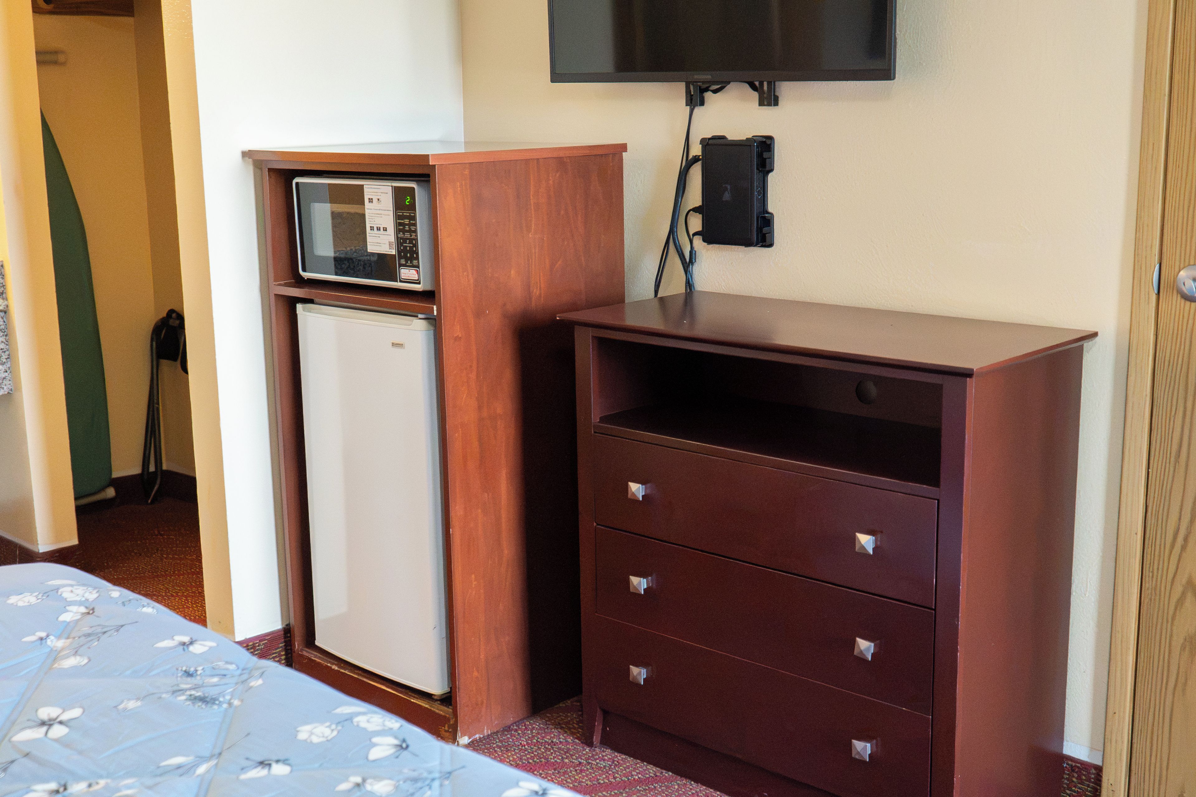 Habitación compacta con cama, un refrigerador pequeño, un aparador de madera con cajones y un televisor montado.