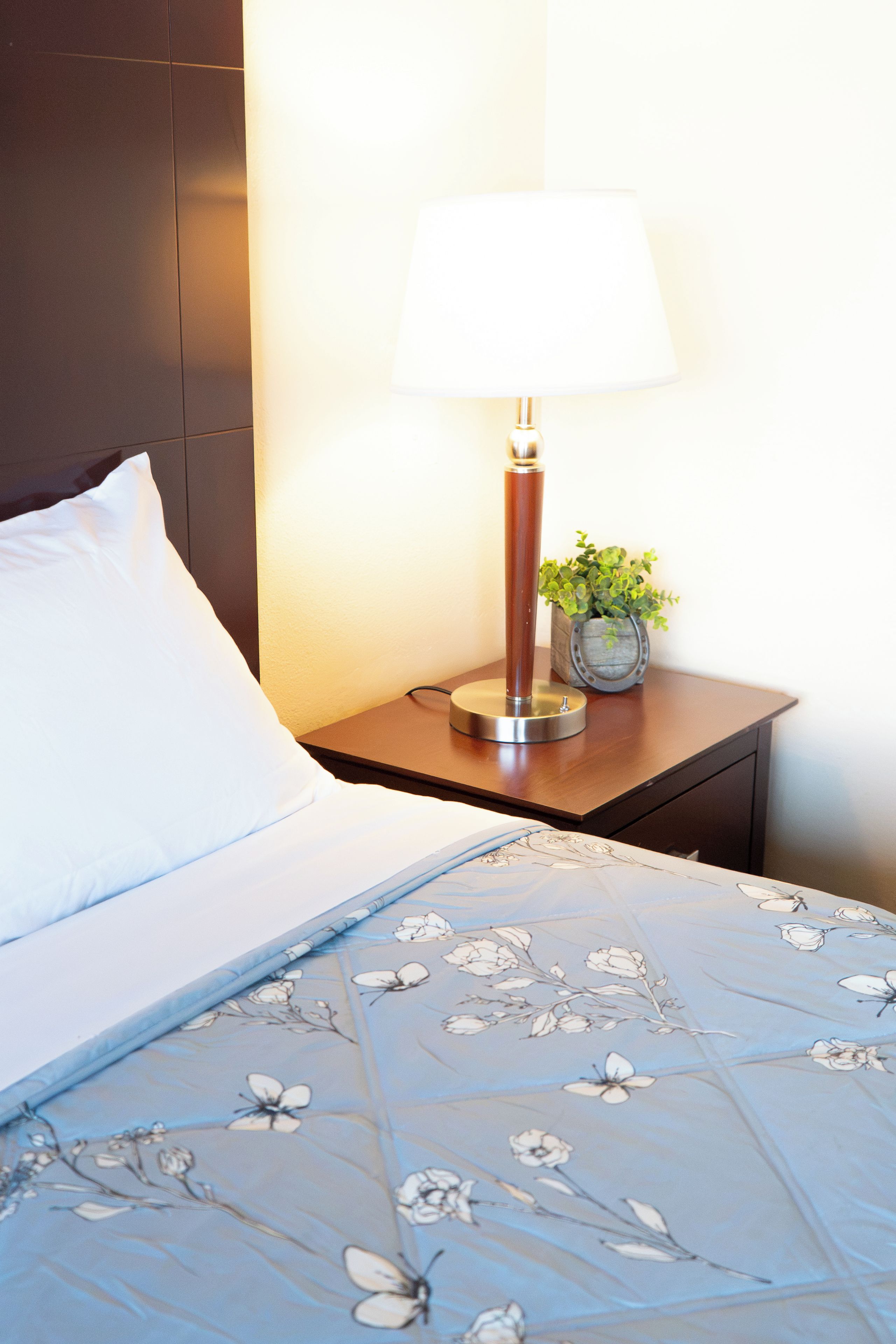 A neatly made bed with a floral-patterned bedspread, a white pillow, and a bedside table with a lamp and a small potted plant.