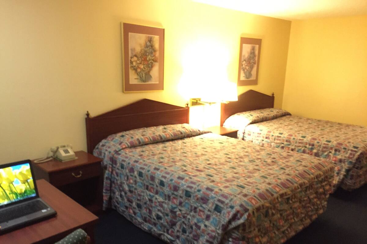 A hotel room with two beds, a desk with a laptop, a phone, and two framed pictures on the wall.