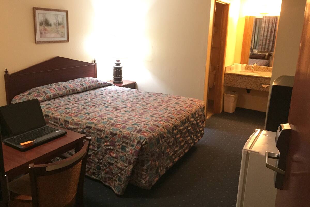A hotel room with a bed, a desk with a laptop, a bedside table with a lamp, a small trash bin, and a refrigerator.