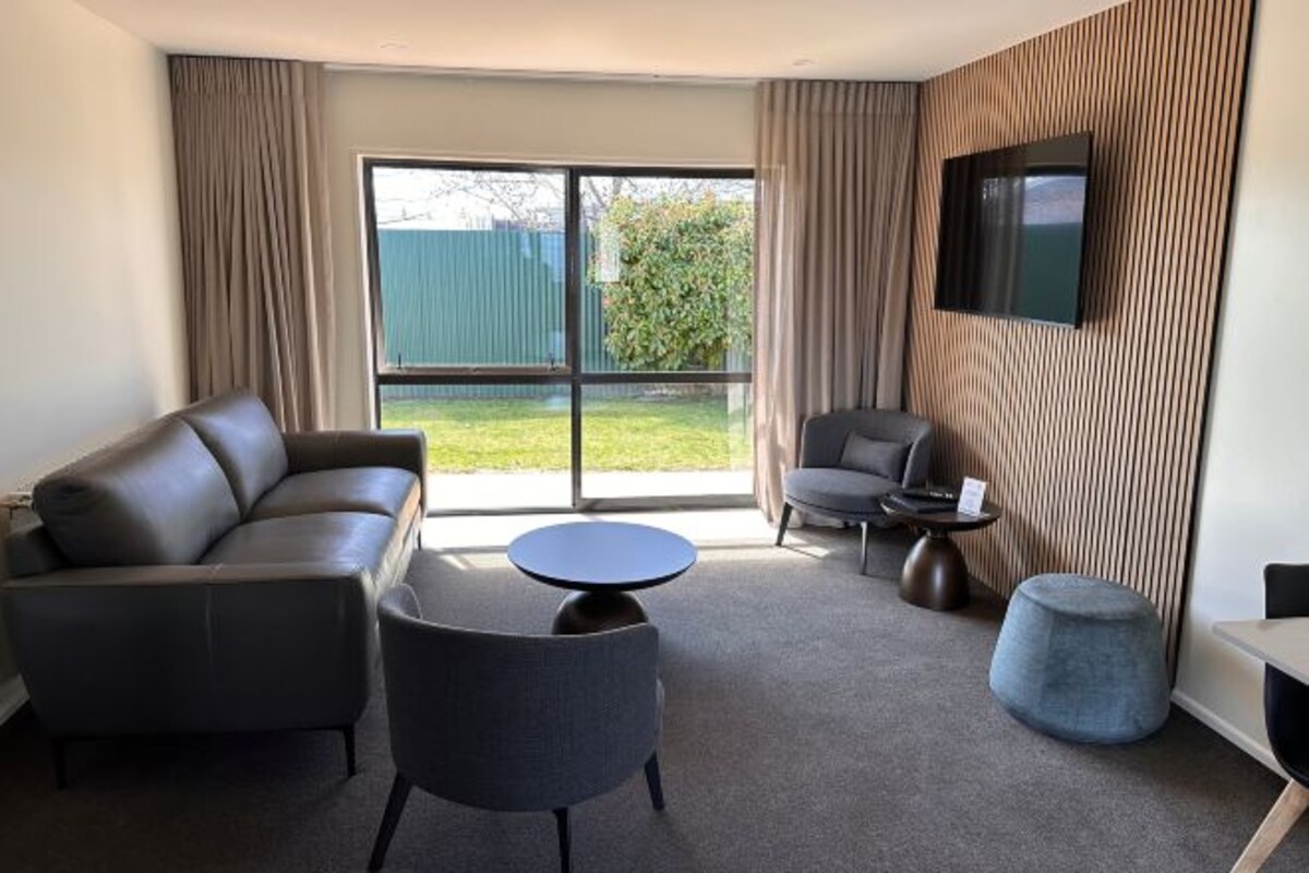 A modern living room with a sofa, armchair, round coffee table, ottoman, and a flat-screen TV mounted on the wall.