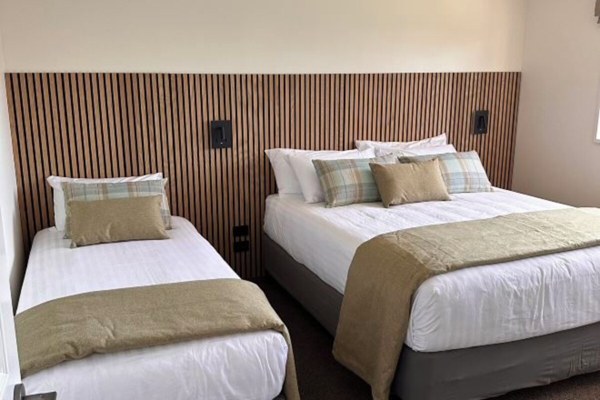 A hotel room with two beds, each with white linens and multiple pillows. The room features a wooden headboard and a neutral color scheme.