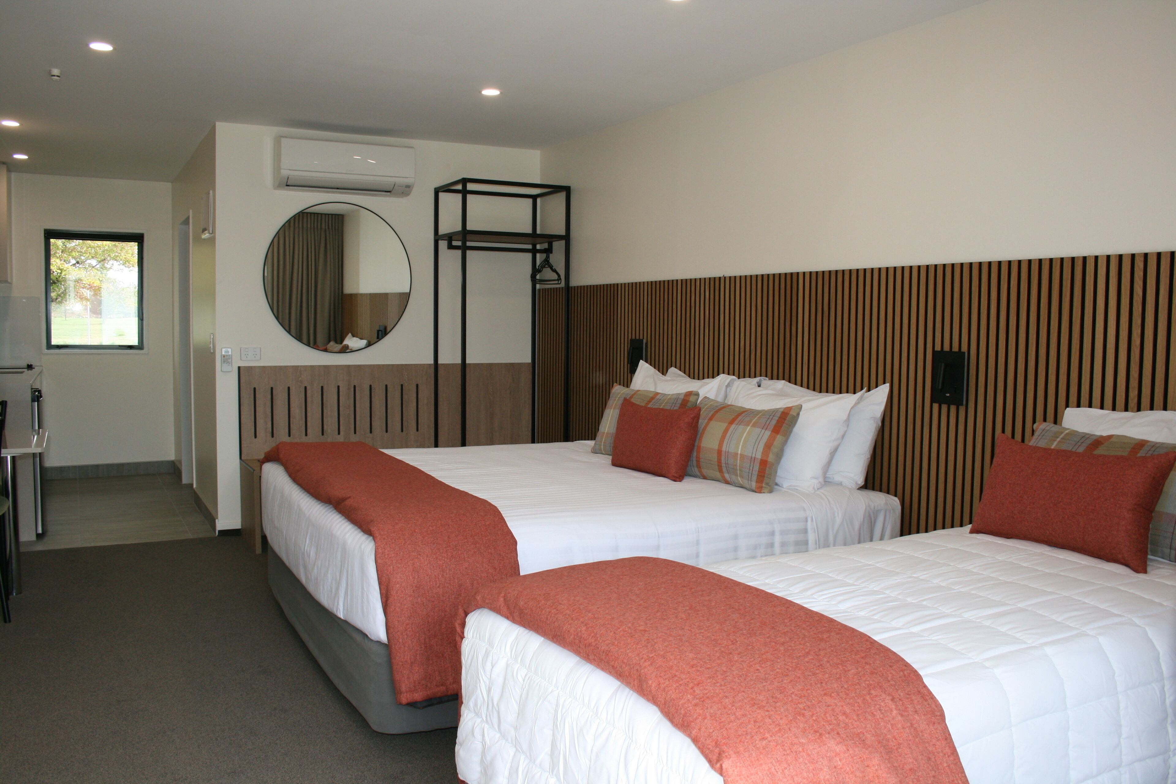 A hotel room with two beds, a round mirror, a wooden headboard, and a window.