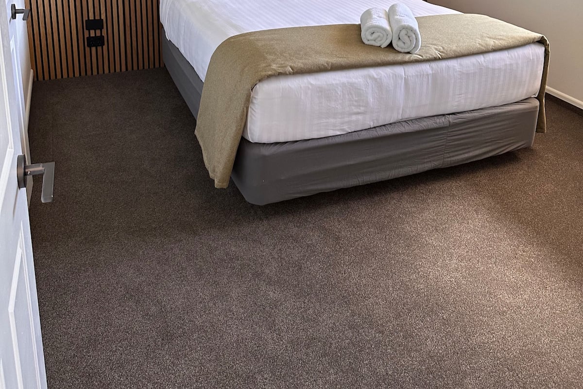 Ein ordentlich bezogenes Bett mit einer braunen Decke und zwei aufgerollten Handtüchern, ein Holterslatten-Kopfteil und ein Fenster mit Blick auf Grünfläche.