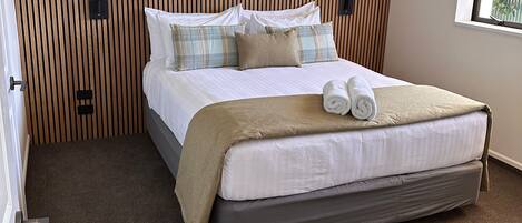 A neatly made bed with a brown blanket and two rolled towels, a wooden slat headboard, and a window with a view of greenery.