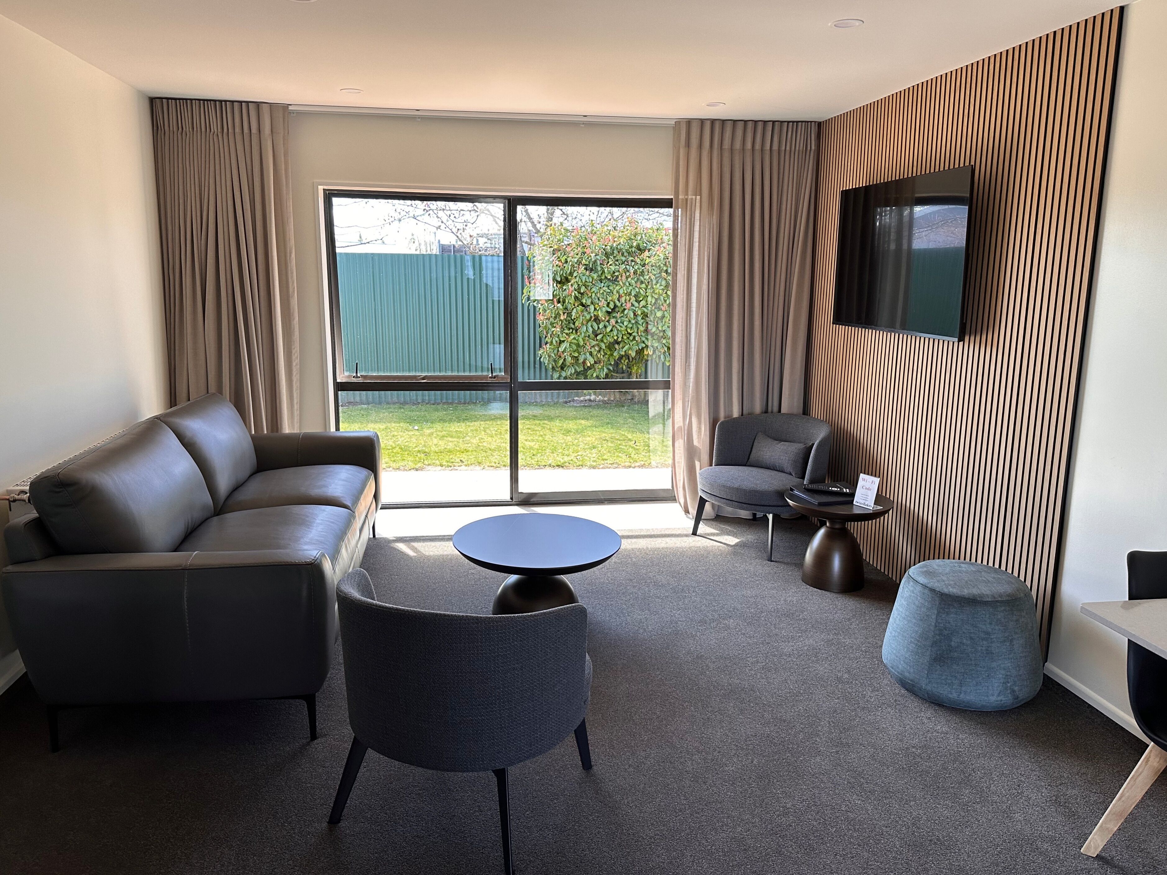 A modern living room with a leather sofa, a grey armchair, a round coffee table, and a small ottoman. A flat-screen TV is mounted on the wall, and the room has large windows with curtains.