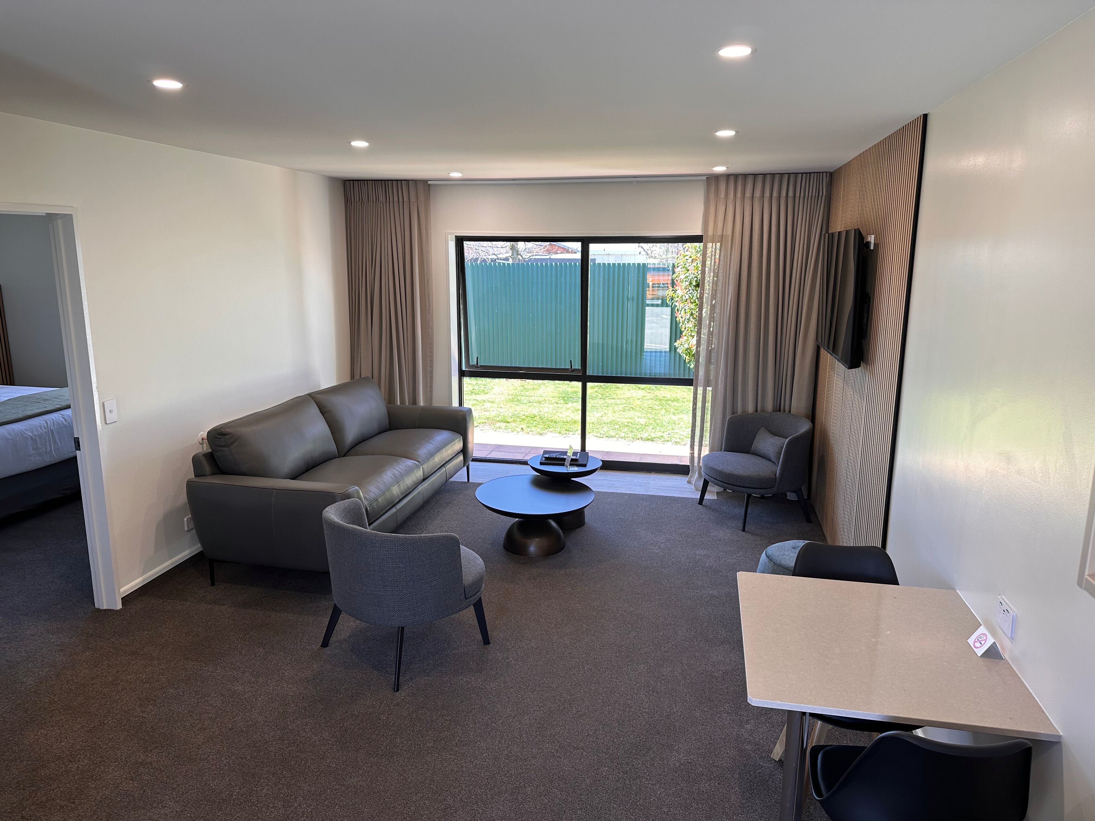 Une pièce moderne et bien éclairée, avec un canapé gris, deux fauteuils, une petite table ronde et un téléviseur fixé au mur.