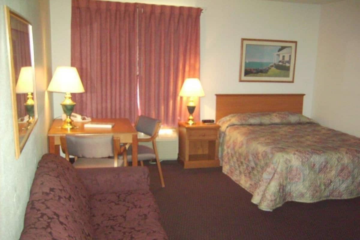 Une chambre d’hôtel comprenant un lit, un canapé, un bureau avec une chaise, deux tables de chevet avec des lampes, un tableau au mur et une fenêtre avec des rideaux.