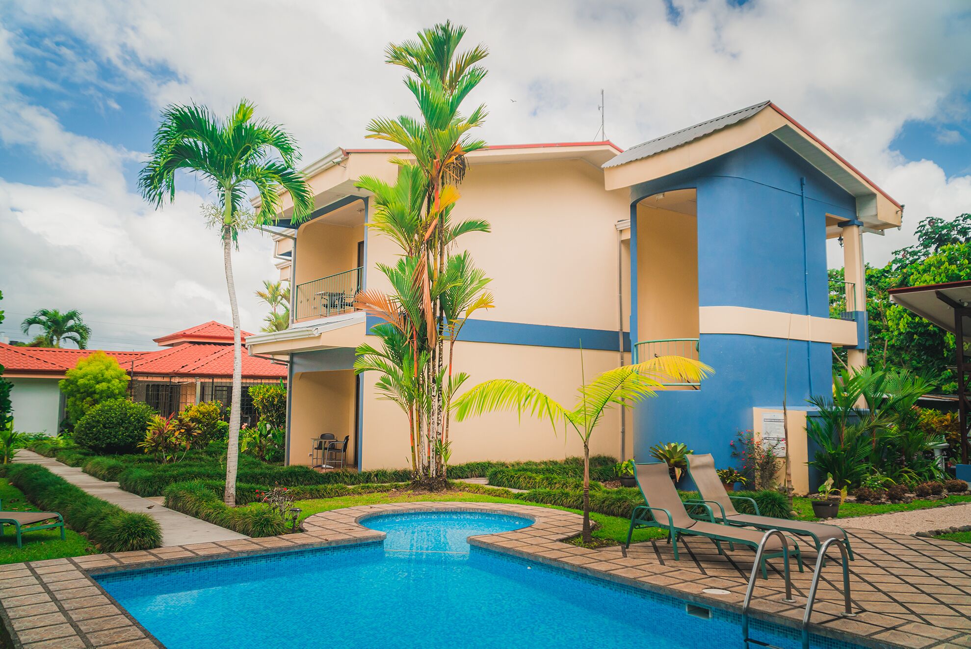 A hotel with a swimming pool, palm trees, and a red-roofed building in the background.