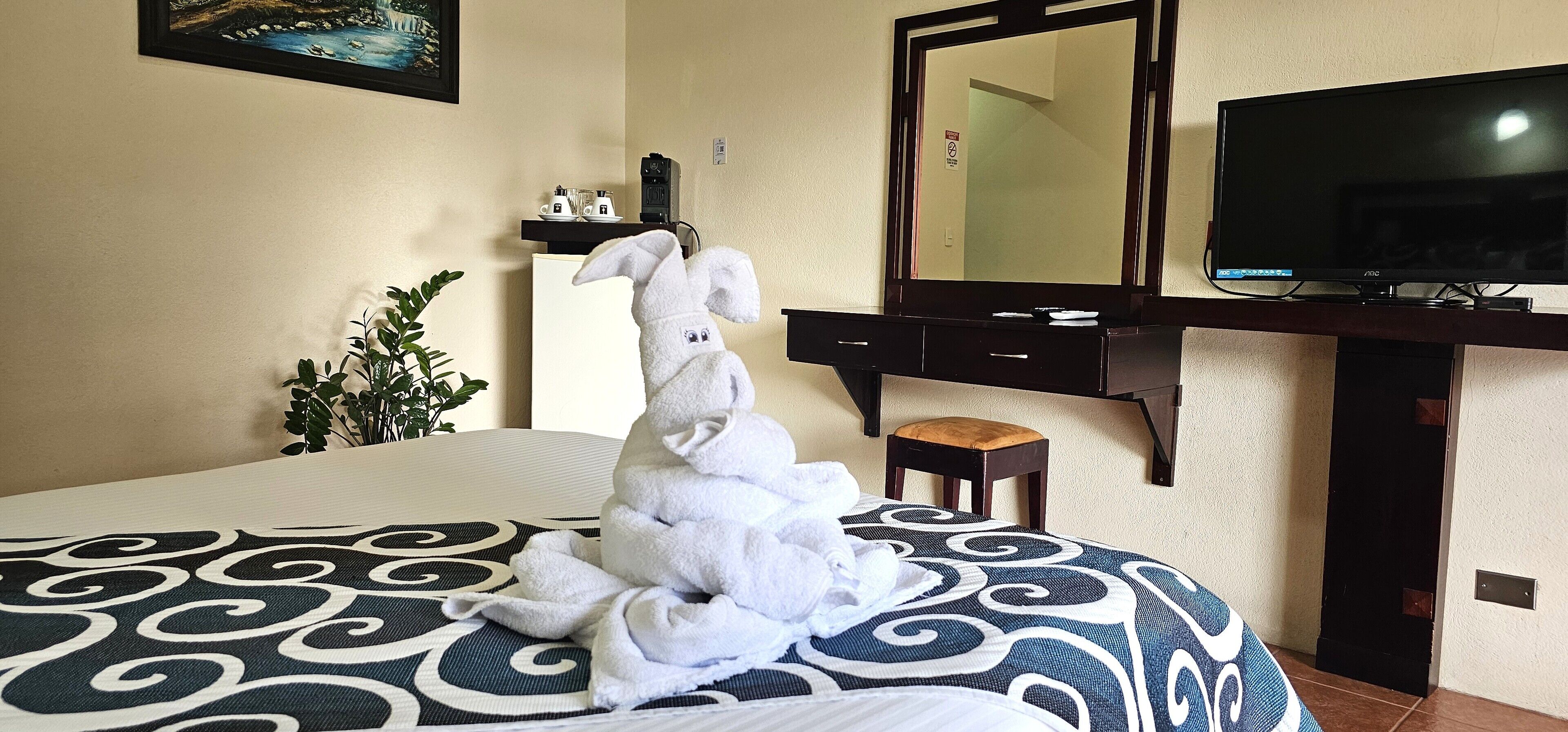 A hotel room with a bed, a television, a desk, a mirror, and a small plant.