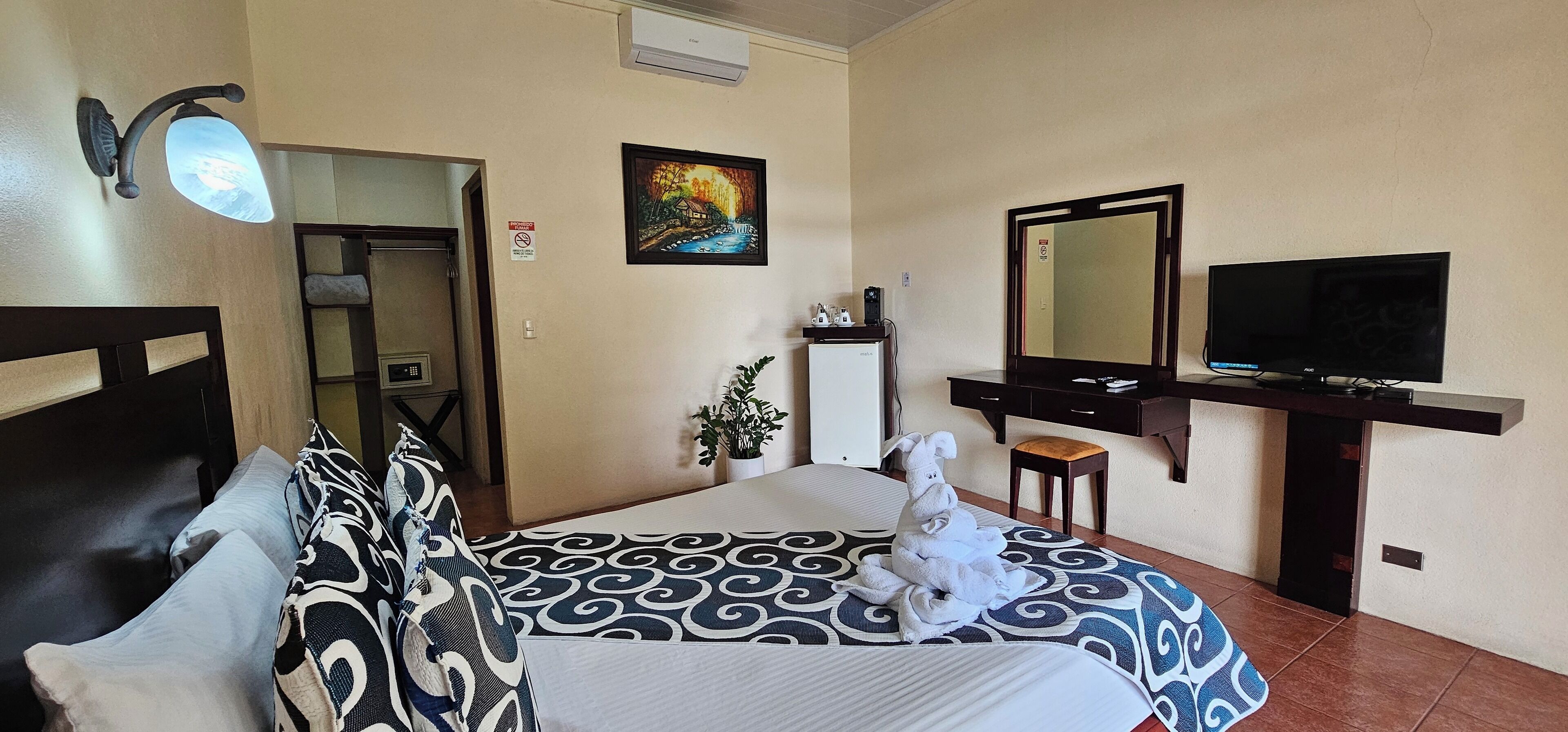 A hotel room with a bed, a TV, a desk, a mirror, a plant, and a painting on the wall.