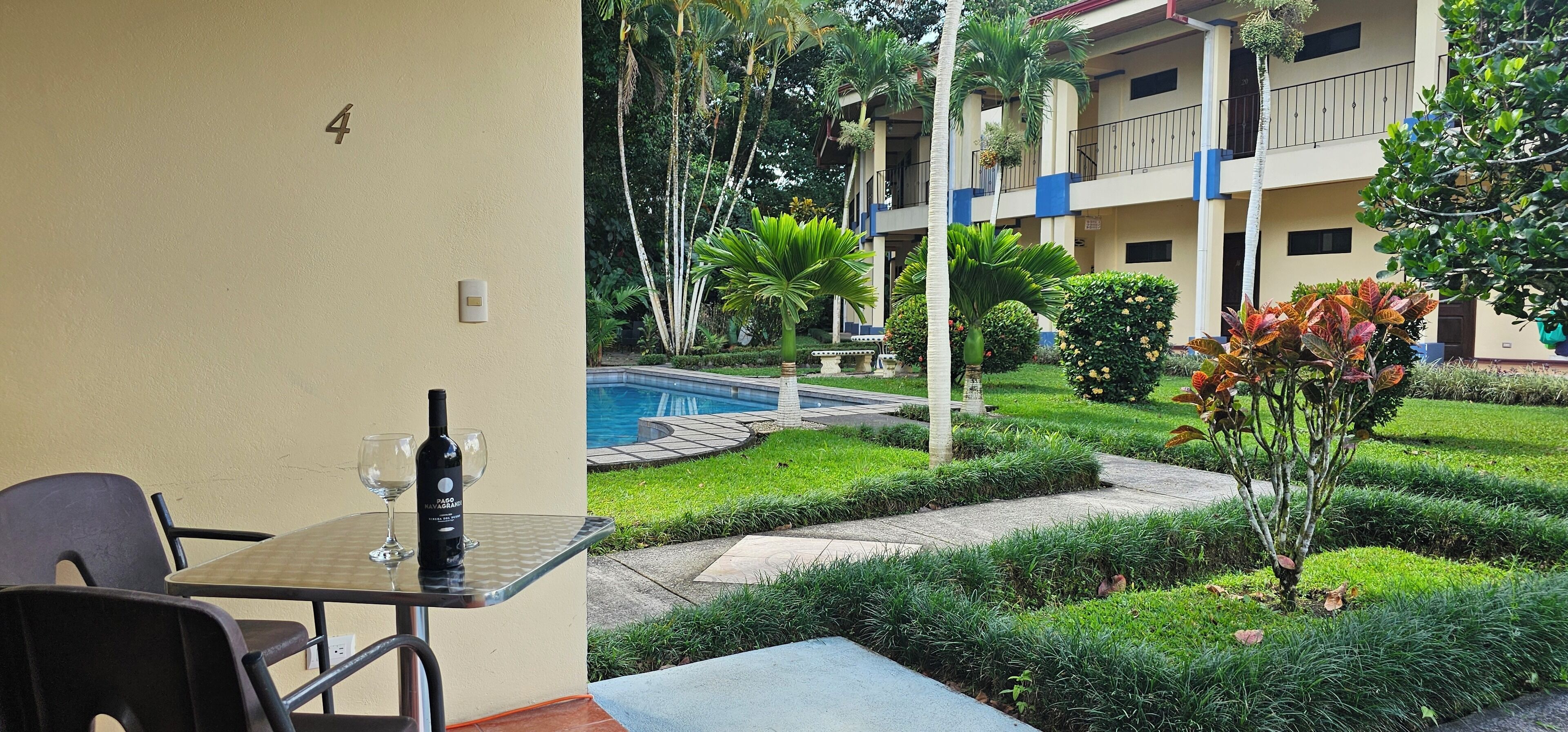 Habitación de hotel con mesa de vidrio, una silla, una botella de vino y vistas a una piscina y jardín.