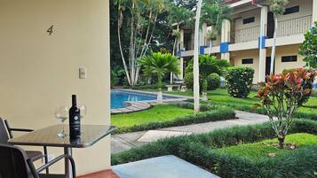 Habitación de hotel con mesa de vidrio, una silla, una botella de vino y vistas a una piscina y jardín.