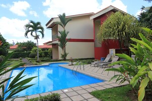 Una piscina con un edificio rojo y color beige al fondo, rodeada de vegetación y un cielo despejado.