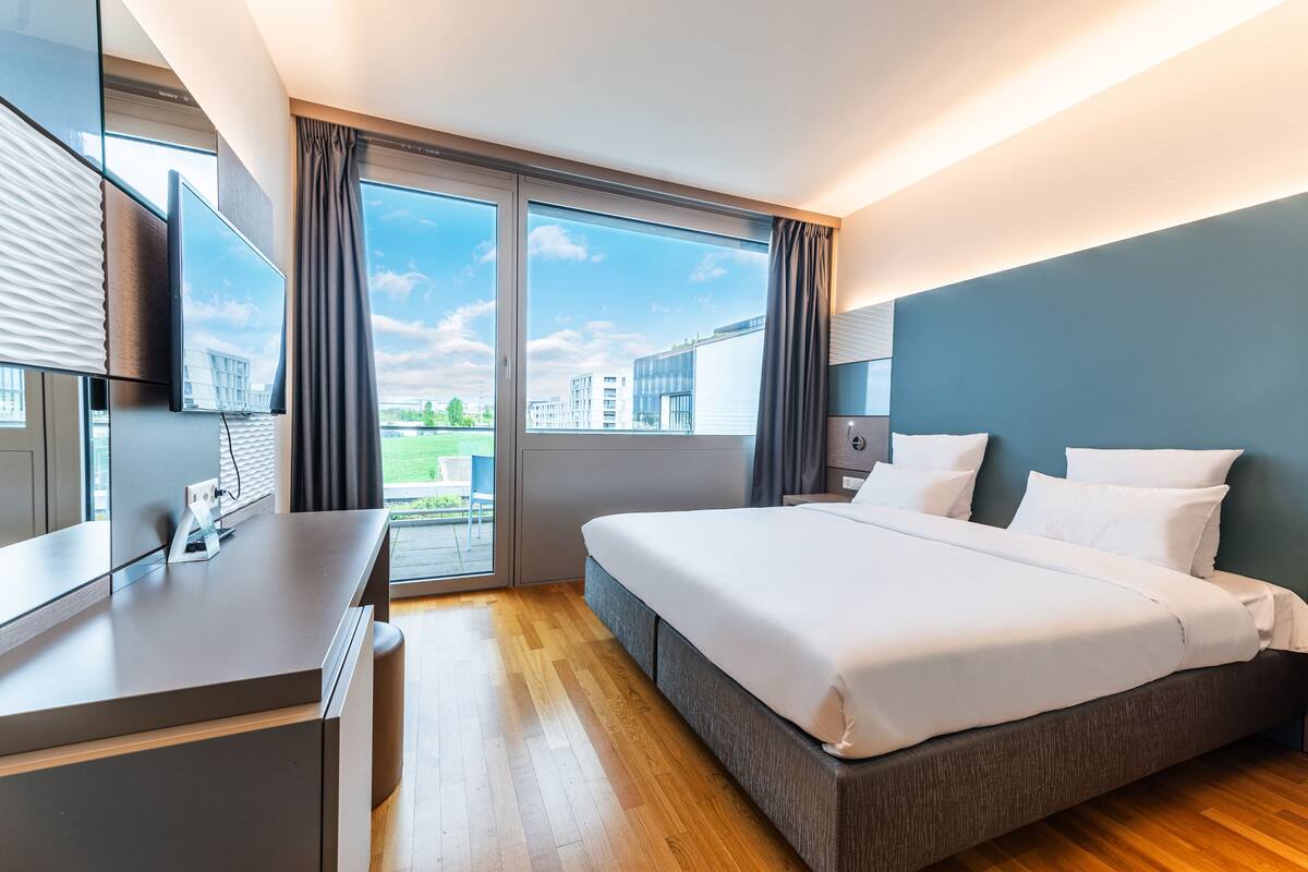 Une chambre d’hôtel moderne dotée d’un grand lit, d’un bureau avec un téléviseur à écran plat et d’une porte-fenêtre coulissante donnant sur un espace extérieur.