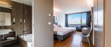 A modern hotel room with a shower, a bed, a desk, and a view of the city.