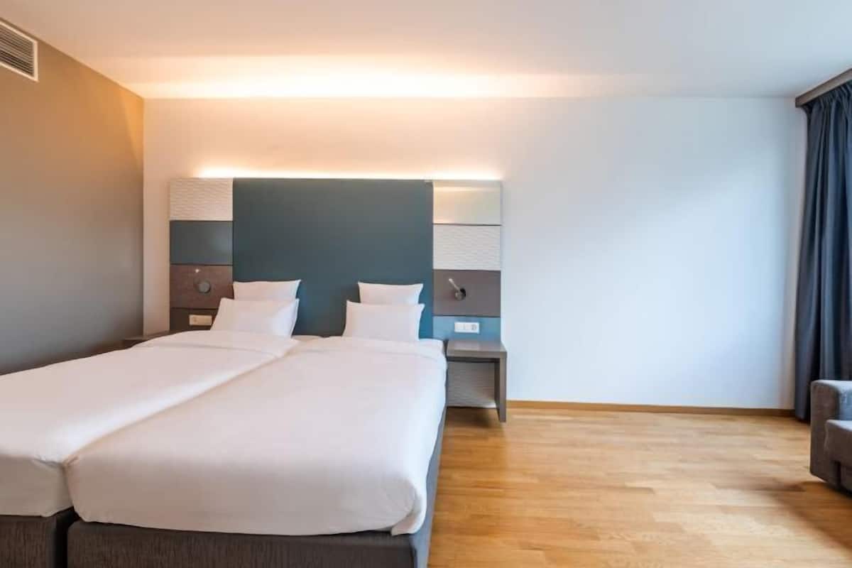 Une chambre d’hôtel avec un grand lit, des tables de chevet, une tête de lit dotée d’un éclairage intégré et une fenêtre avec des rideaux.