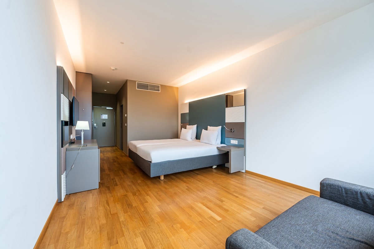 Une chambre d’hôtel moderne avec un plancher en bois, un lit recouvert de draps blancs, un canapé gris, un bureau avec une lampe et un grand miroir au mur.