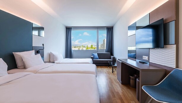 Een moderne hotelkamer met twee bedden, een bureau, een stoel, een televisie en een groot raam met uitzicht op de stad.