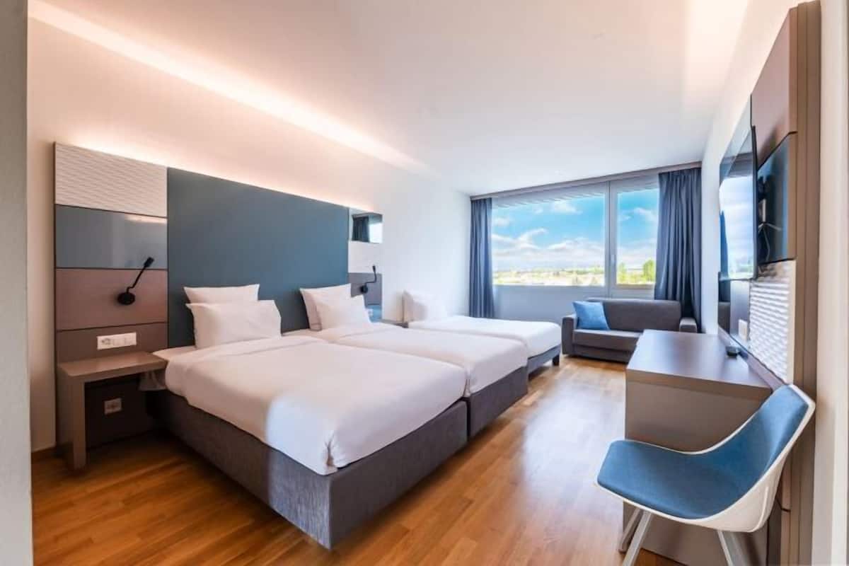 Une chambre d’hôtel avec deux lits, un bureau, une chaise, une télévision et une grande fenêtre donnant sur la ville.