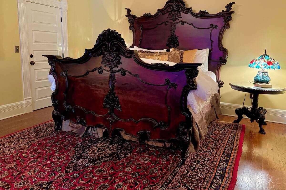 A room with a large, ornate bed with a dark headboard, a red and black patterned rug, a small table with a lamp, and a white door.