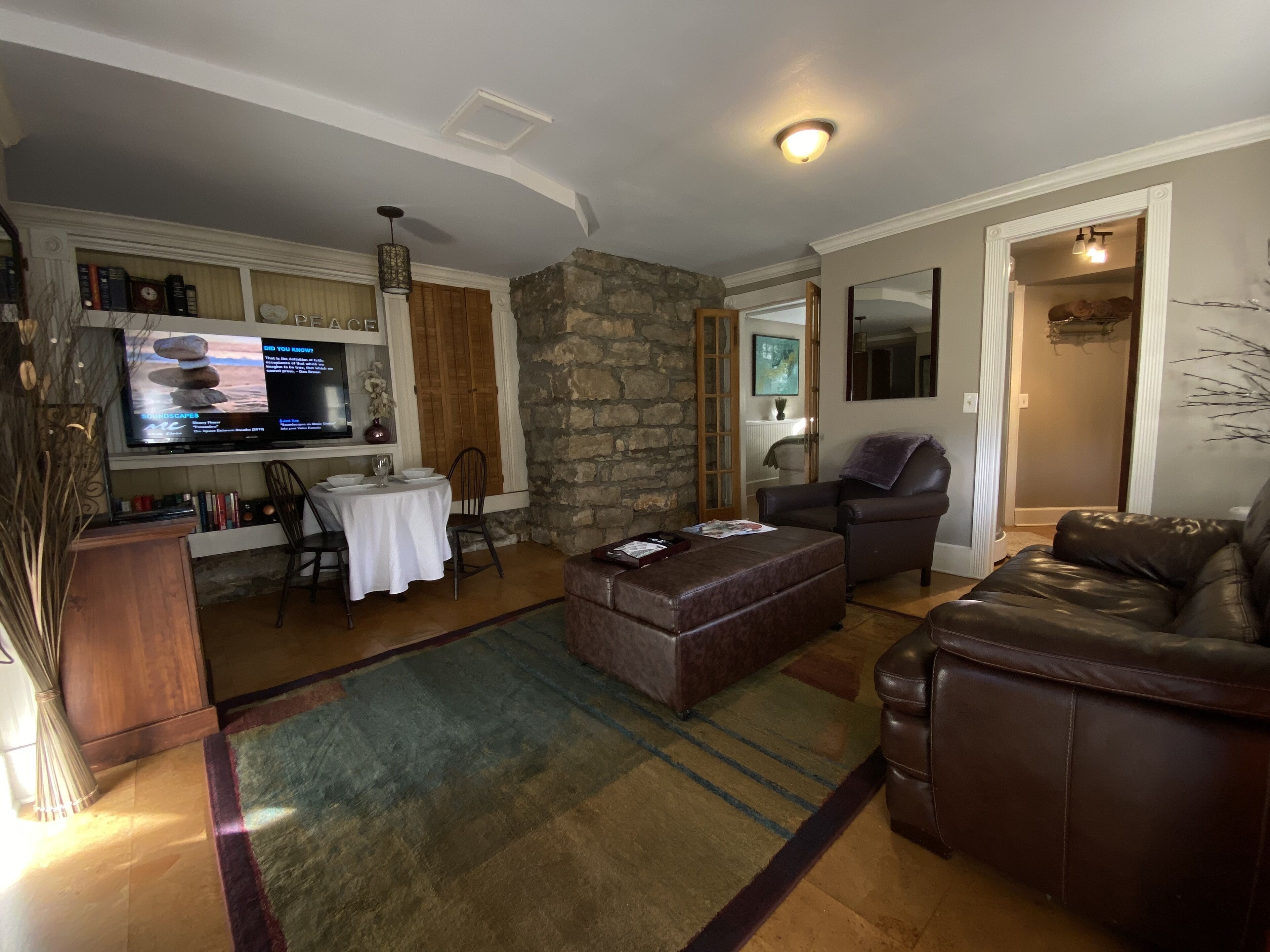 A living room with a stone accent wall, a television, a dining table with chairs, a brown leather sofa, and a patterned rug.