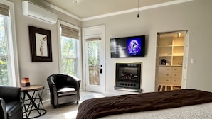 A bedroom with a large bed, a fireplace, a TV mounted on the wall, and a chair by the window. - Eureka Zen Cottages (Eureka Springs)
