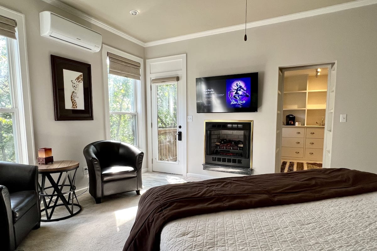 A bedroom with a large bed, a fireplace, a TV mounted on the wall, and a chair by the window.