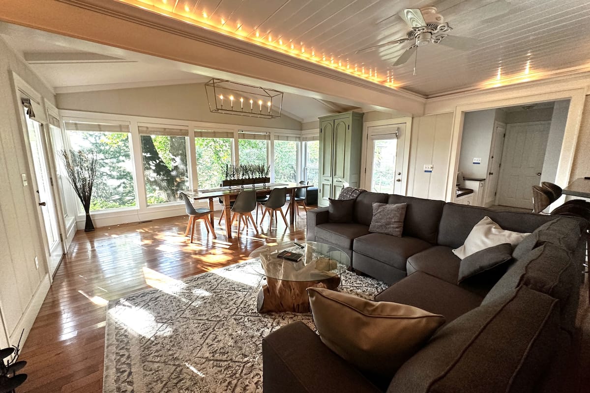 A spacious living room with a large sofa, a coffee table, and a dining area with a wooden table and chairs. There is a ceiling fan and a large window providing natural light.
