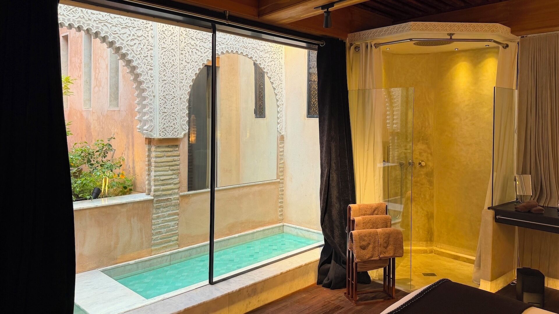 A bathroom with a glass shower enclosure, a small seating area with a towel, and a view of a courtyard with a pool.
