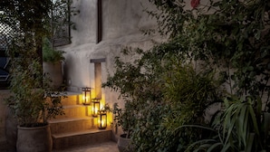 Une cour avec des marches menant à une porte, entourée de plantes en pot et illuminée par des lanternes.