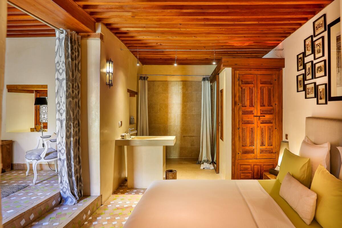 Ein Hotelzimmer mit Holzboden, einem Bett mit grün-weißer Bettwäsche, einem Badezimmer mit Waschbecken und Spiegel sowie einer Holztür.
