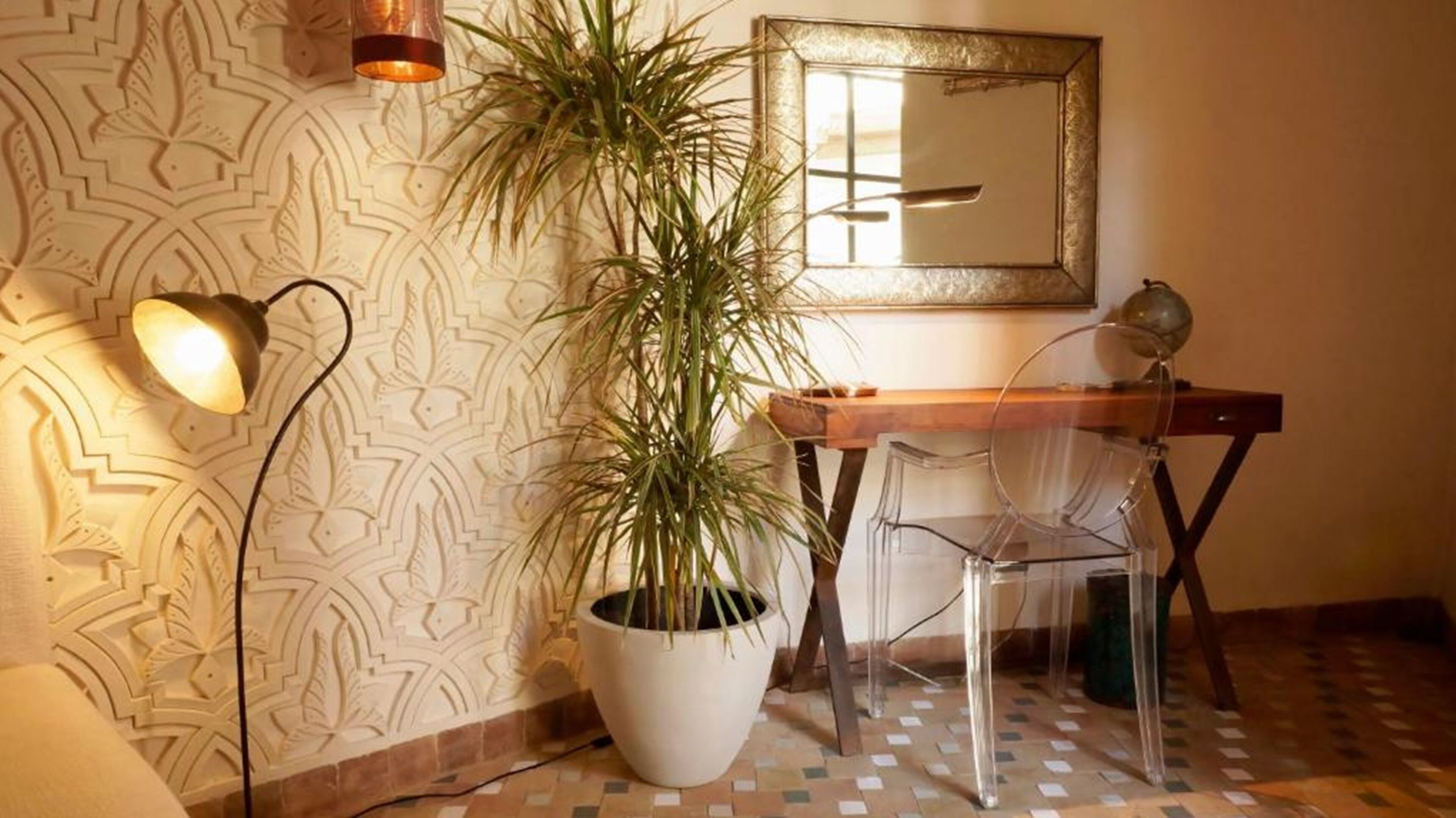 A room with patterned wallpaper, a potted plant, a desk with a chair, and a wall-mounted mirror.