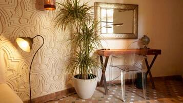 A room with patterned wallpaper, a potted plant, a desk with a chair, and a wall-mounted mirror.