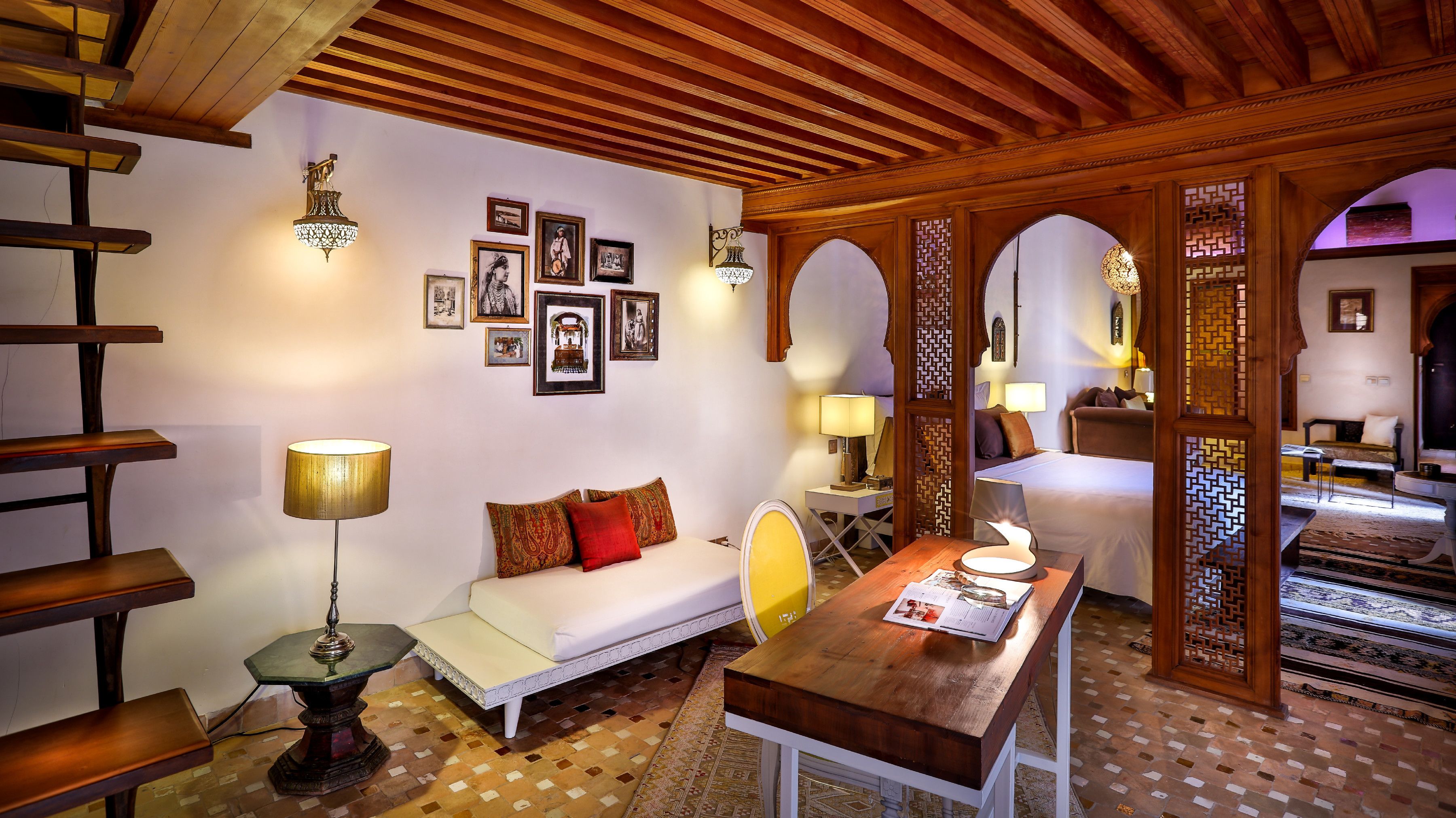 A cozy room with a wooden ceiling, a sofa with patterned cushions, a small table with a lamp, and a wall adorned with framed pictures.