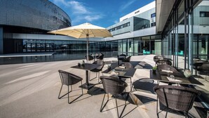 An outdoor seating area with tables and chairs, a large umbrella, and a modern building in the background. - Aquatis Hotel (Lausanne)