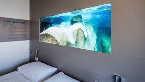 A hotel room with a bed, a wall-mounted TV displaying an underwater scene, and a sign with information about Switzerland. - Aquatis Hotel (Lausanne)