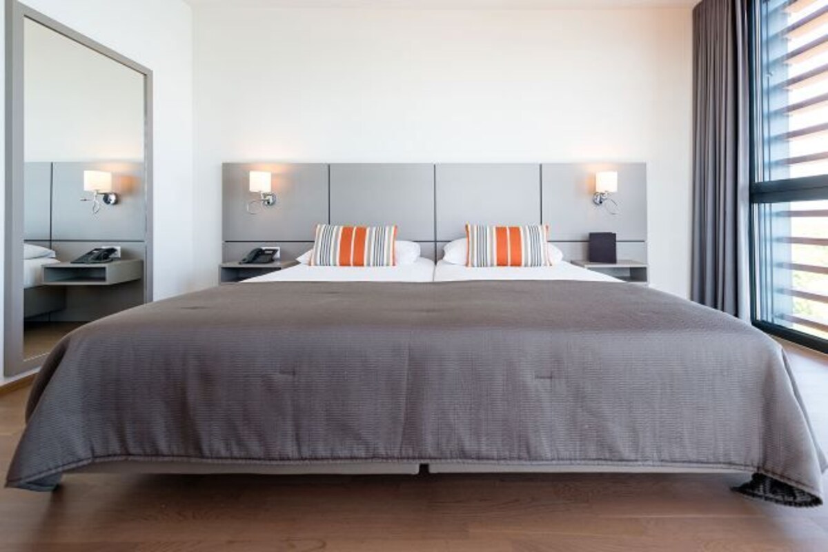 A hotel room with a large bed, two bedside tables with lamps, a mirror, and a window with blinds.