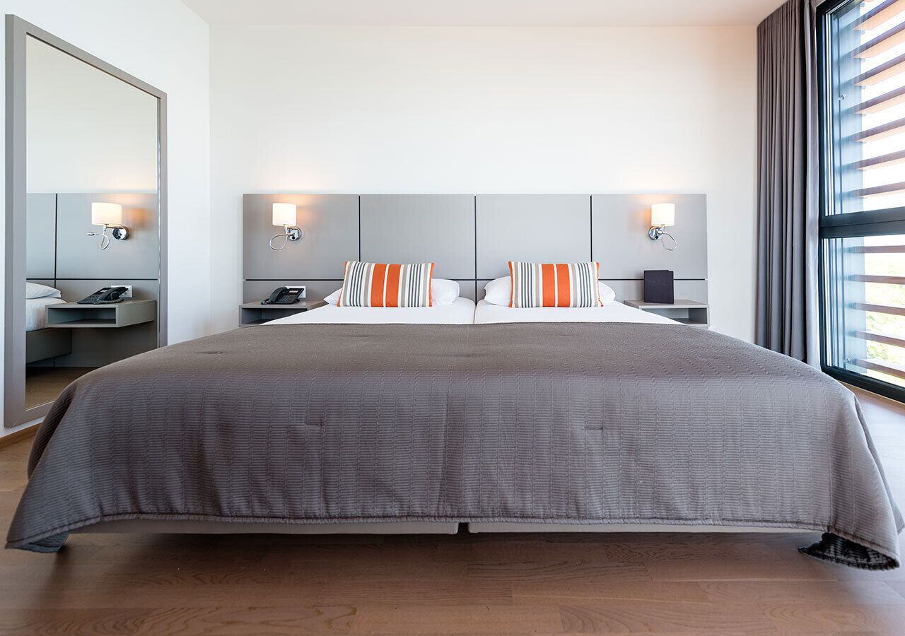 A modern hotel room with a large bed, two bedside tables, a mirror, and a window with blinds.