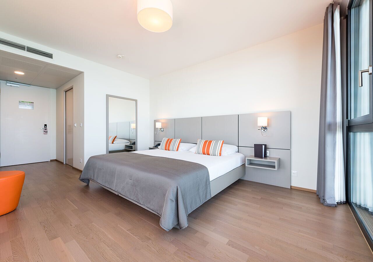 A modern hotel room with a large bed, bedside tables, a nightstand, and a window with curtains.