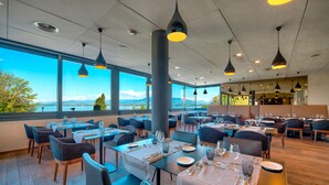 A restaurant with tables set for dining, large windows offering a view of the outdoors, and hanging pendant lights. - Lake Geneva Hotel (Versoix)