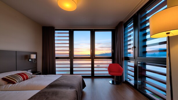 A hotel room with a large bed, a red chair, and a view of the sunset through floor-to-ceiling windows. - Lake Geneva Hotel (Versoix)
