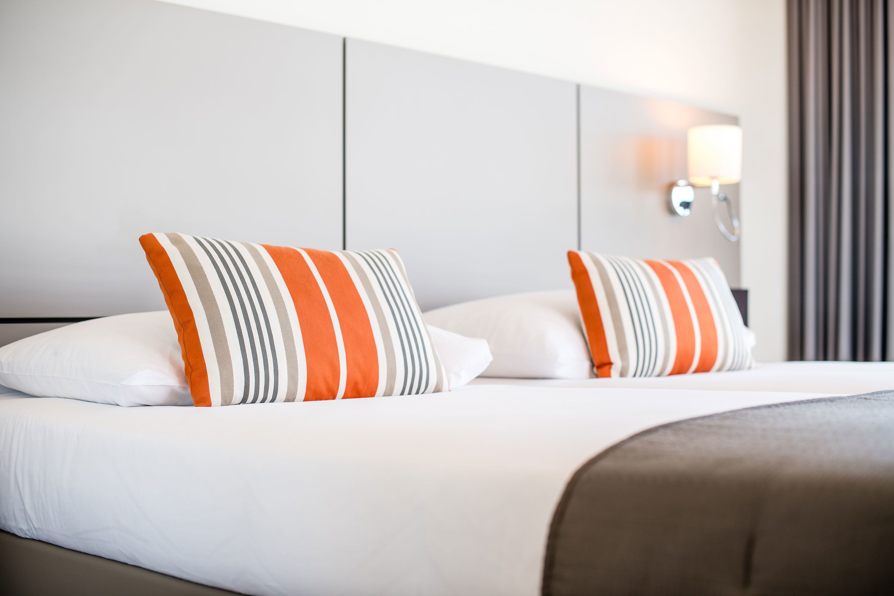A neatly made bed with white and striped pillows, a grey headboard, and a wall-mounted light fixture.