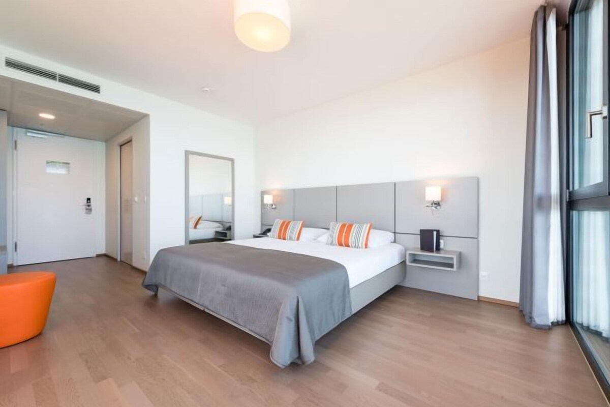 A modern hotel room with a large bed, a bedside table, a nightstand, and a window with curtains.