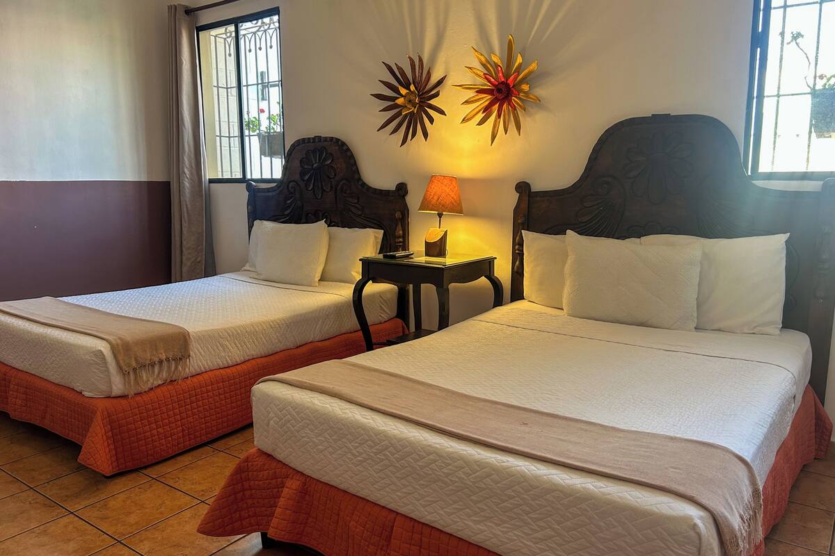 Dos camas individuales con sábanas blancas, una mesita de noche con lámpara, decoración en la pared y una ventana con barrotes.