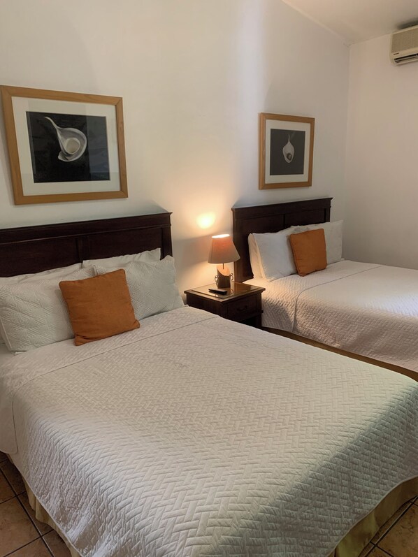 A hotel room with two beds, each with white bedding and a brown pillow. There are framed pictures on the wall above the beds, and a nightstand with a lamp between the beds.