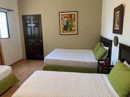 A hotel room with two beds, a dark wooden door, a painting on the wall, and a small table with a plant.